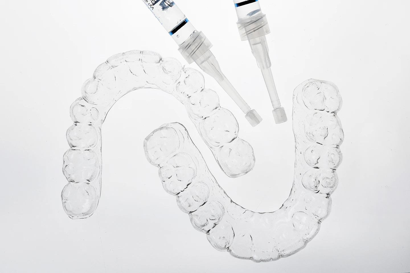 Two clear dental aligner trays with two syringes of whitening gel on a white background.
