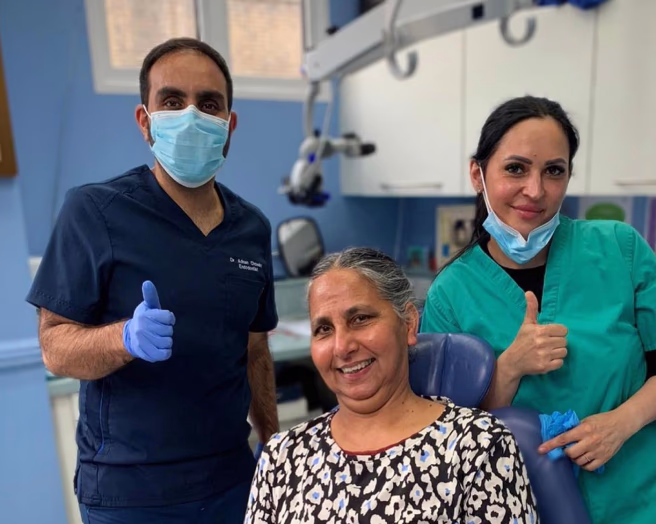 Smiling woman seated in a dental chair with two healthcare professionals in masks and scrubs giving thumbs up behind her in a dental clinic.