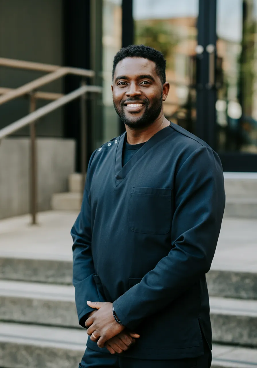 Smiling man in dark medical scrubs standing outside near stairway and glass doors.