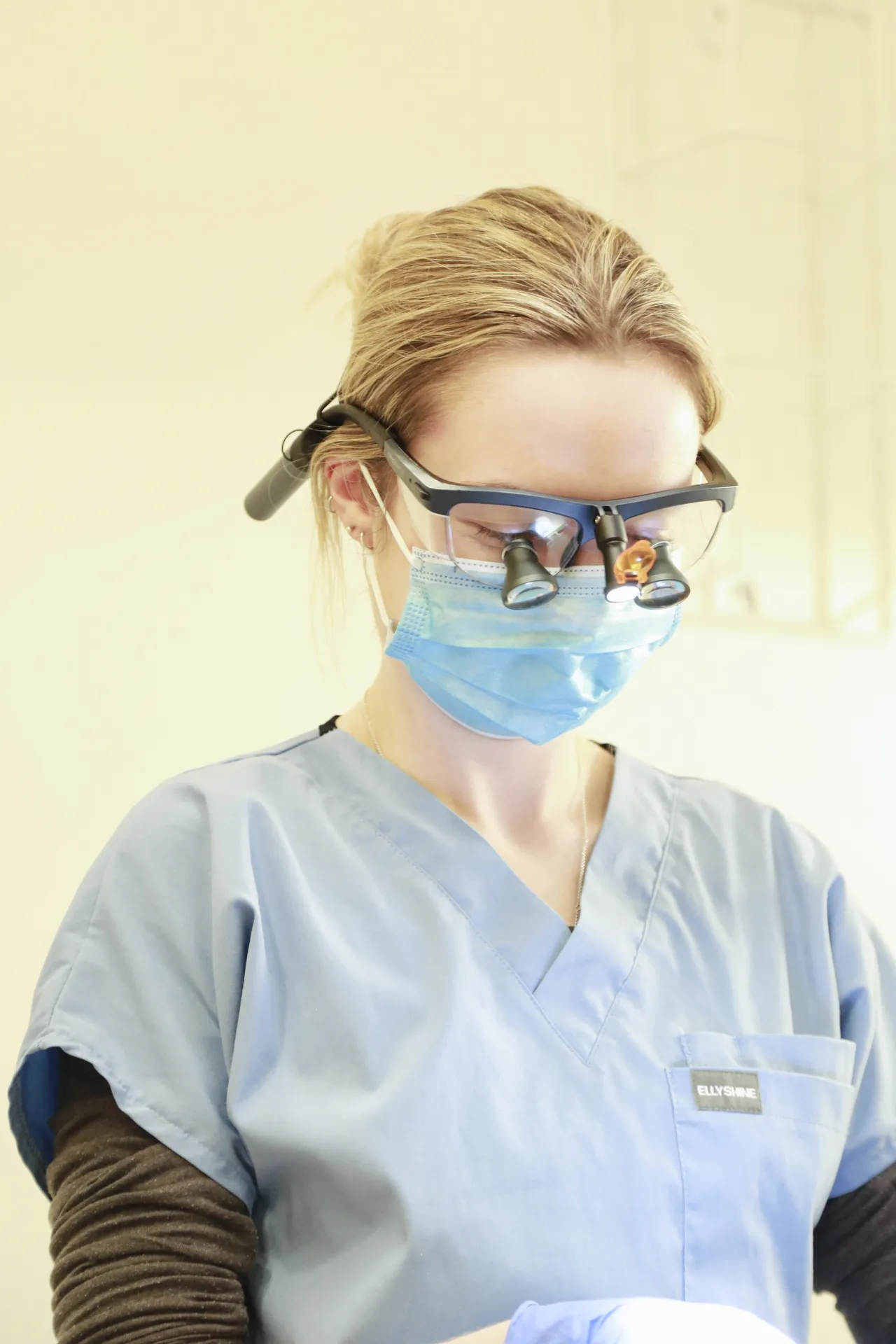 Female healthcare professional wearing surgical scrubs, mask, and magnifying glasses with a light.