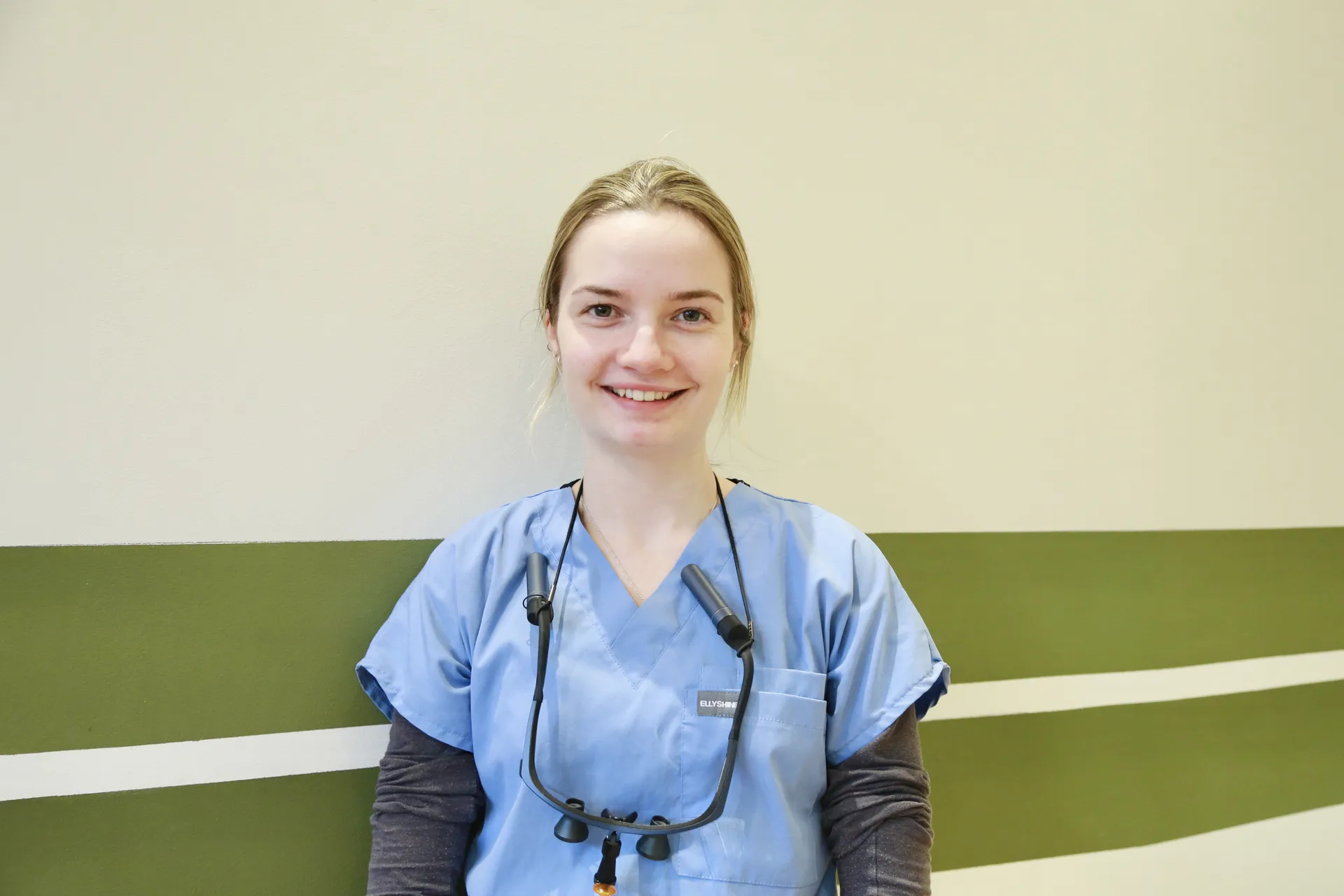 Smiling female healthcare worker in blue scrubs and dark long sleeves against a beige and green wall.
