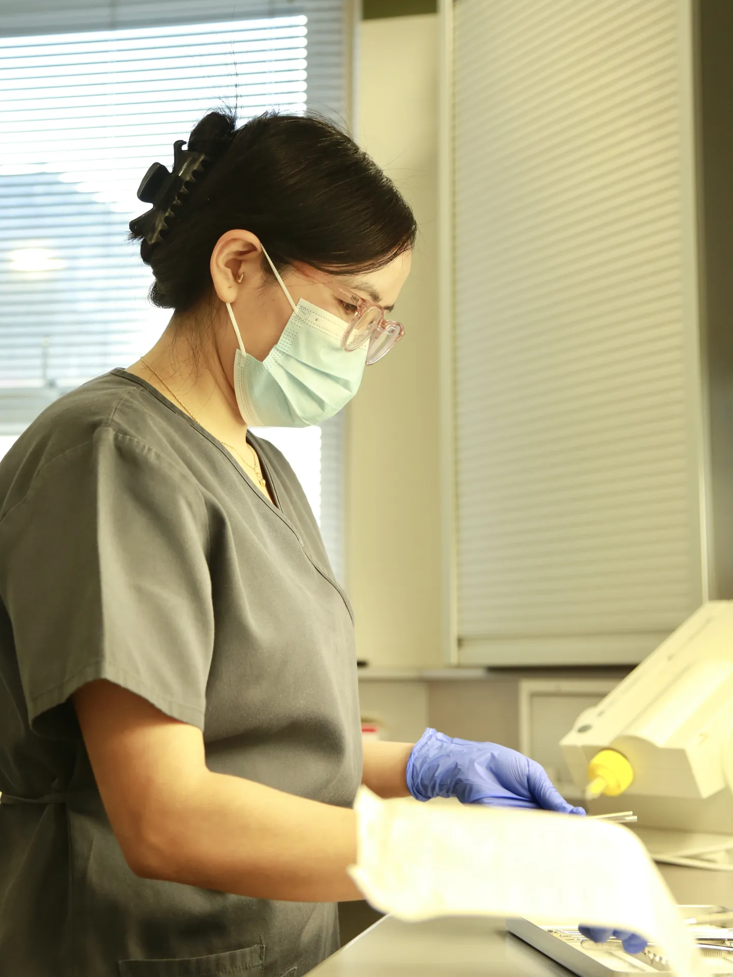 Woman in medical scrubs, mask, and gloves working with instruments in a clinical setting.
