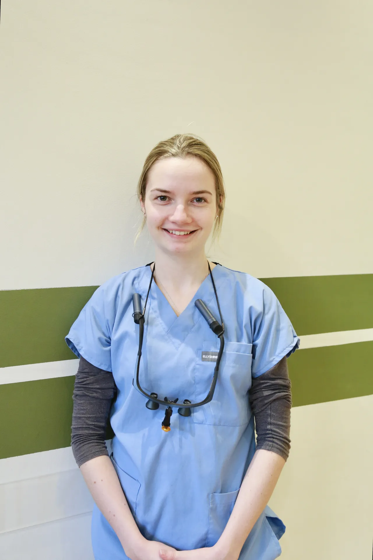 Smiling woman in blue medical scrubs with a dental loupe around her neck against a beige wall.