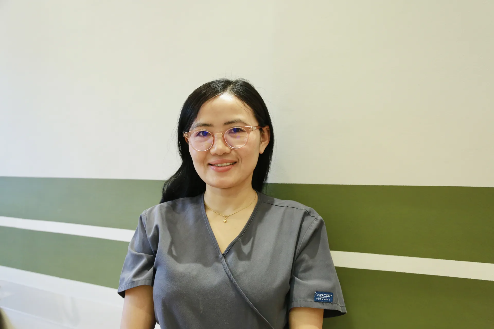 Smiling woman in gray medical scrubs with glasses standing against green and white wall.