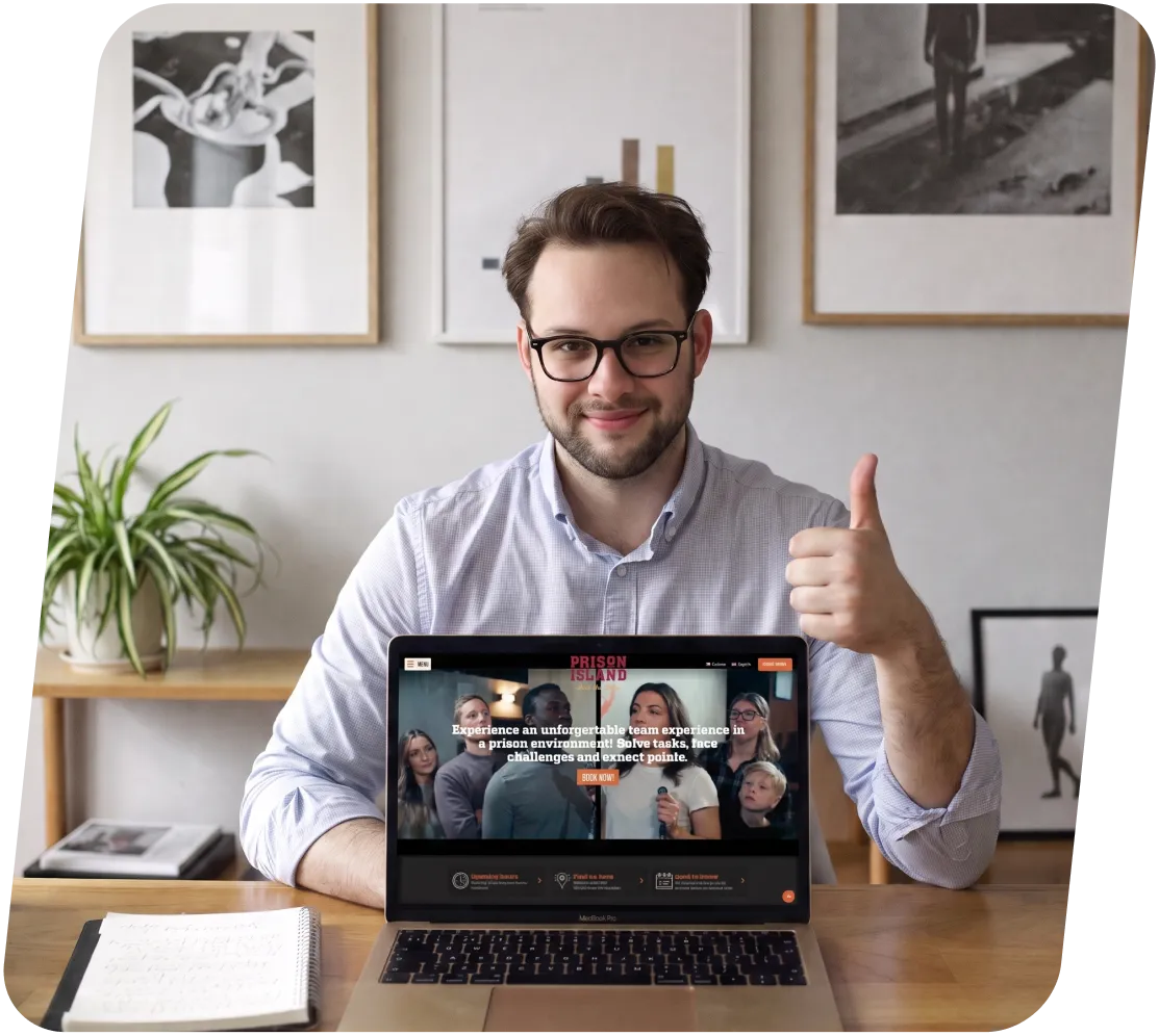 Man with glasses sitting at a desk showing laptop screen and giving thumbs up.