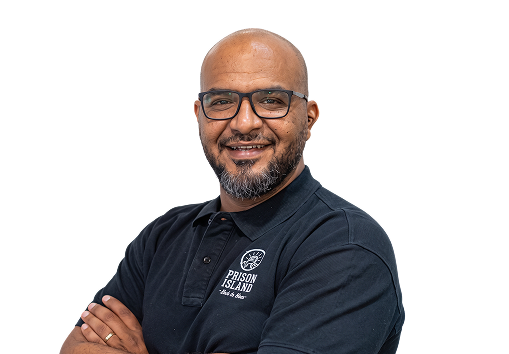Smiling man with glasses and beard wearing a black Prison Island polo shirt with arms crossed.