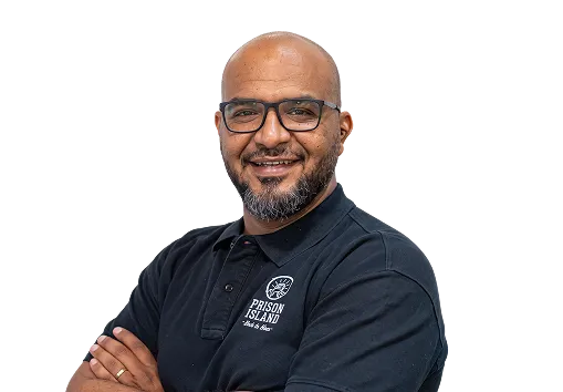 Smiling man with glasses and beard wearing a black Prison Island polo shirt with arms crossed.