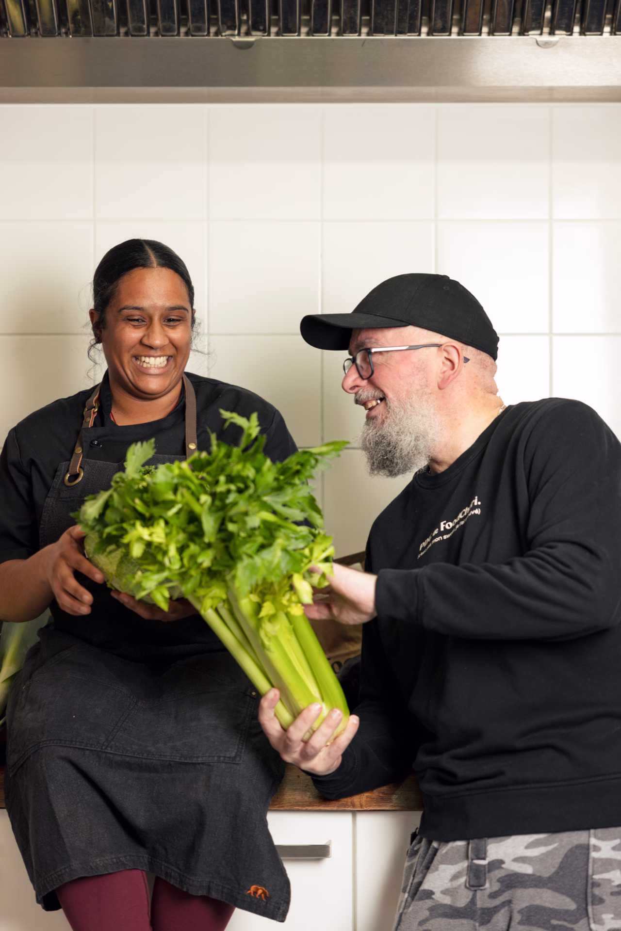 Deux cuisiniers souriants tenant une botte de céleri dans une cuisine blanche.