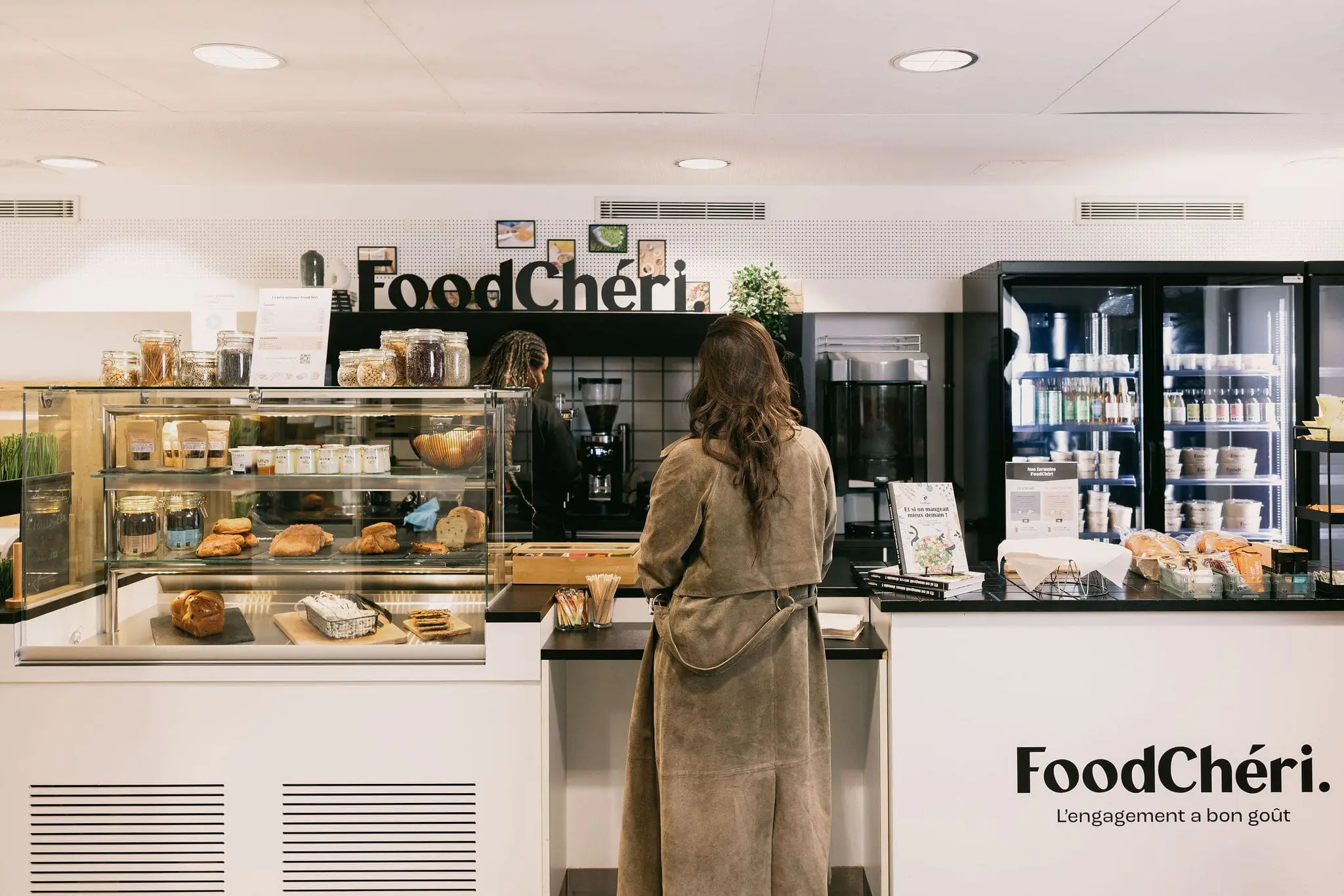 Client en manteau long face au comptoir d’une boutique FoodChéri avec des pâtisseries et boissons exposées.
