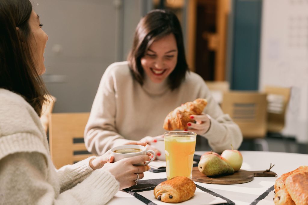 Deux femmes souriantes prenant le petit déjeuner avec croissants, café, jus d'orange, pommes et poire.