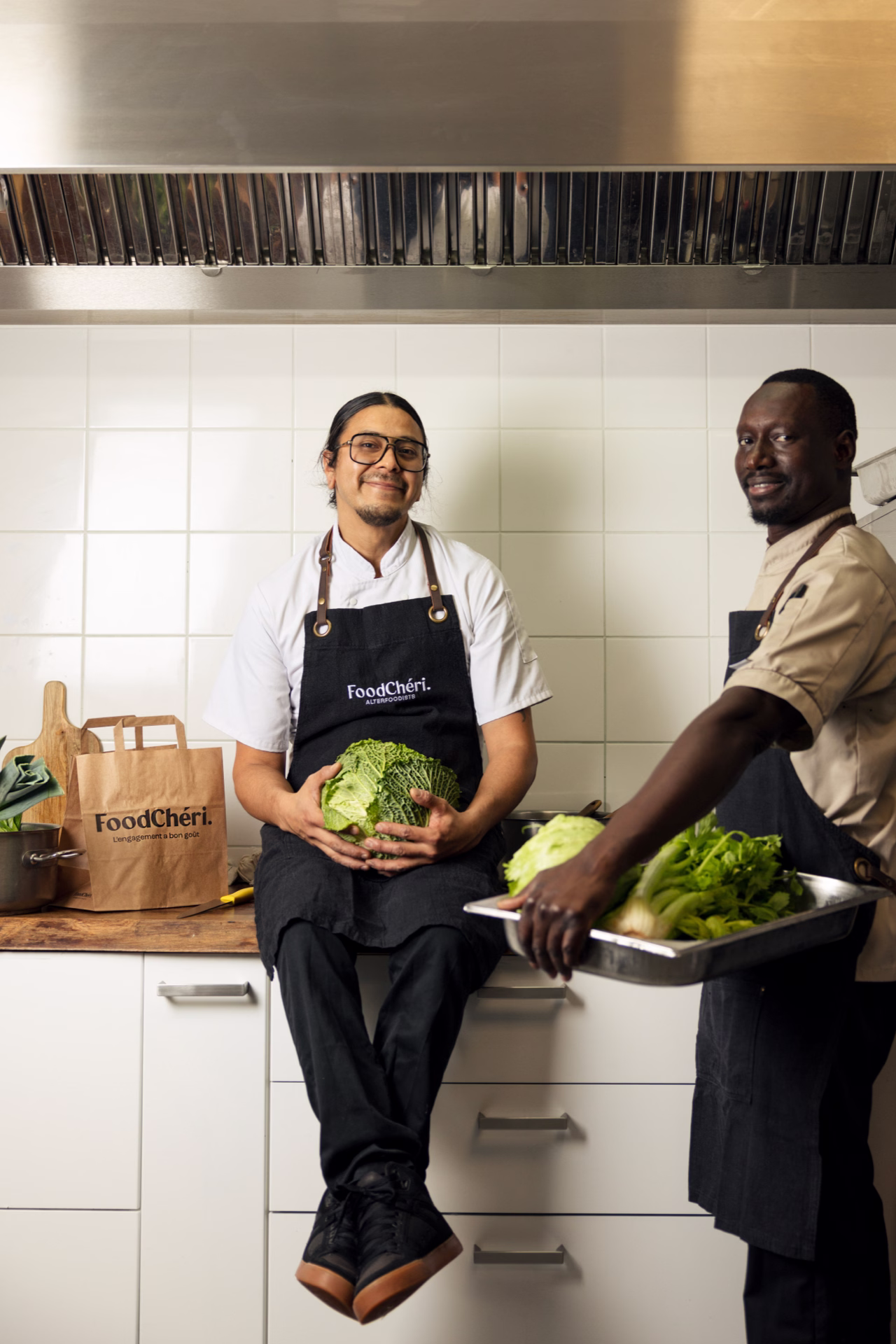 Deux chefs souriants en tabliers FoodChéri avec légumes verts dans une cuisine professionnelle.