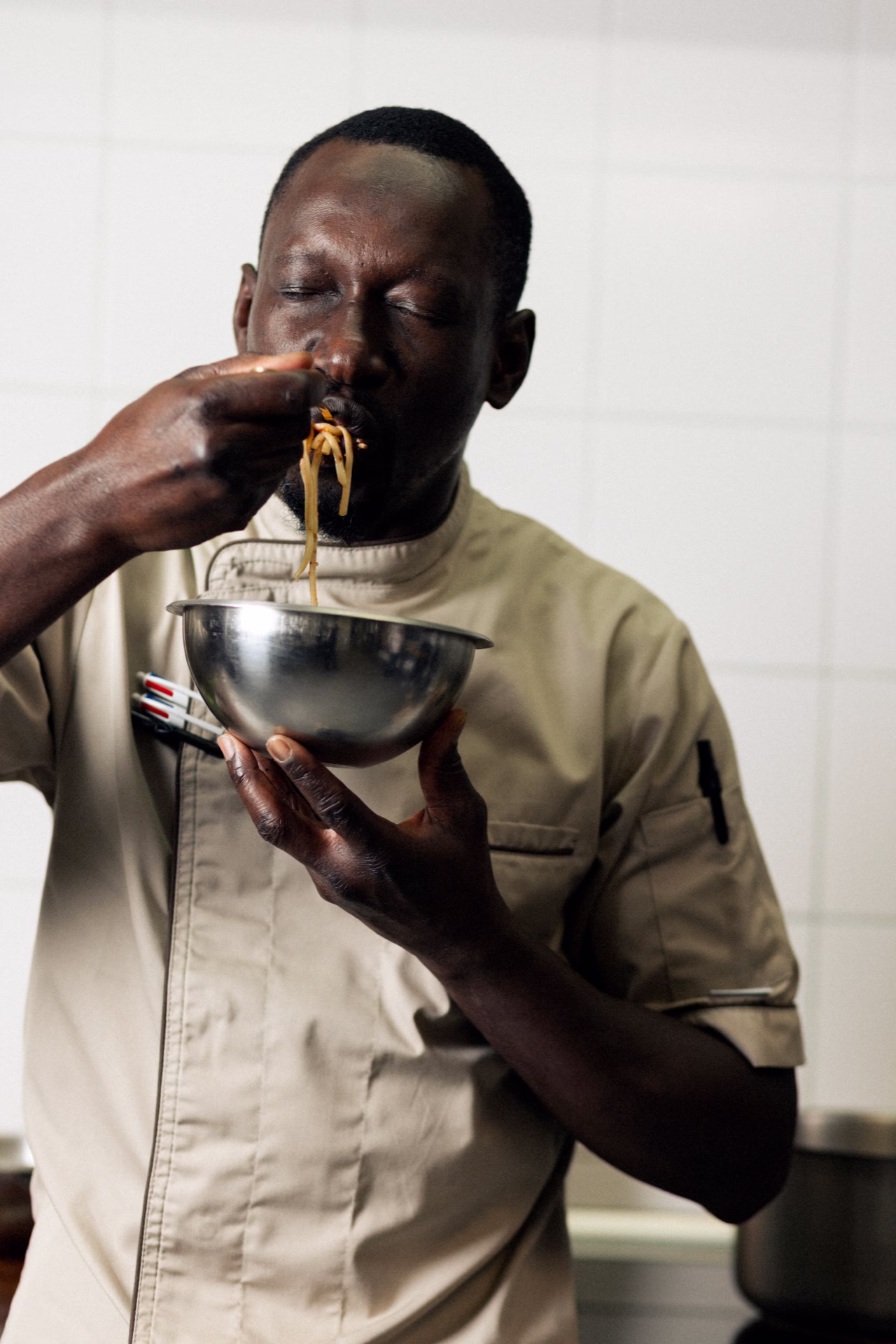 Chef en blouse beige dégustant des pâtes dans un bol en acier inoxydable dans une cuisine.