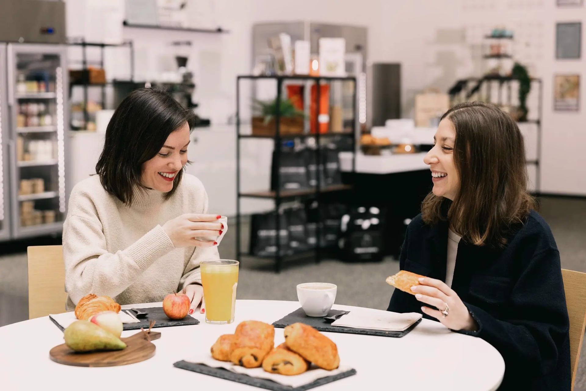 Deux femmes souriantes prennent un petit-déjeuner avec jus, café, fruits et viennoiseries.