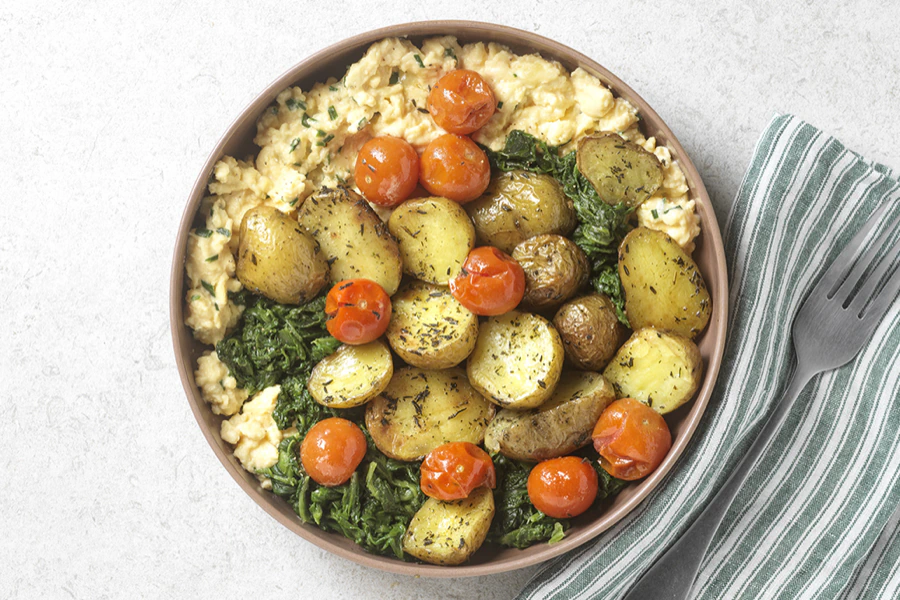 Bol de pommes de terre rôties, tomates cerises, épinards et œufs brouillés à côté d'une fourchette.