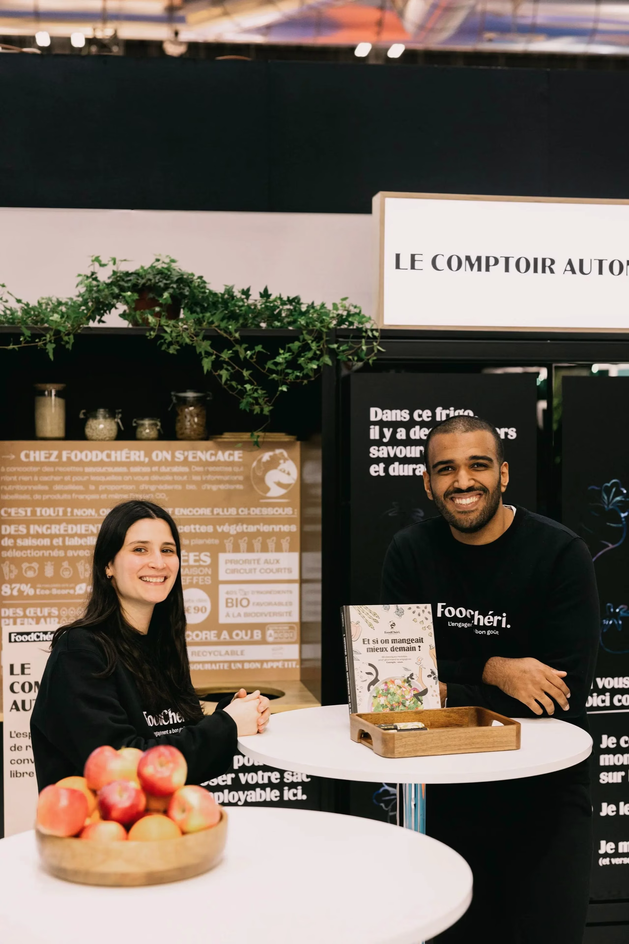 Deux personnes souriantes devant un stand FoodChéri avec pommes, plantes et affiches explicatives.