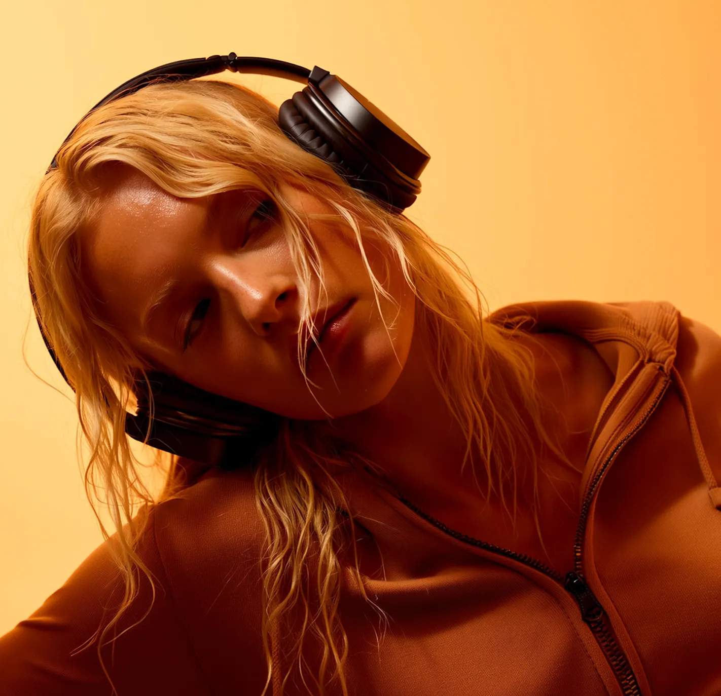 Blonde woman wearing black headphones and a brown zip-up hoodie against a warm orange background.