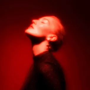 Portrait of a person with short blonde hair and closed eyes, head tilted back, illuminated by red light.
