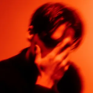Blurred image of a person covering their face with their hand against an orange background.