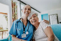 Smiling nurse in blue scrubs embracing and sitting with an elderly woman on a couch in a bright room.