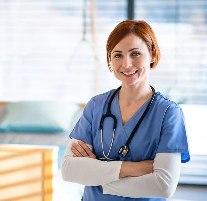 Smiling female healthcare professional in blue scrubs with stethoscope around her neck, arms crossed.