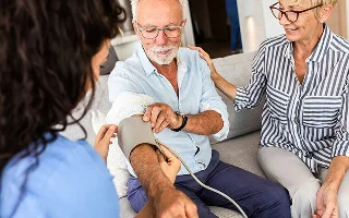 Older man having his blood pressure measured by a healthcare professional while a woman supports him on the couch.