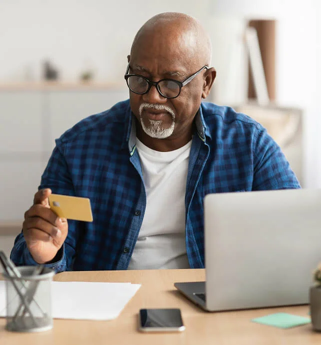 Older man wearing glasses and a blue checkered shirt holding a credit card while looking at a laptop screen at a desk.