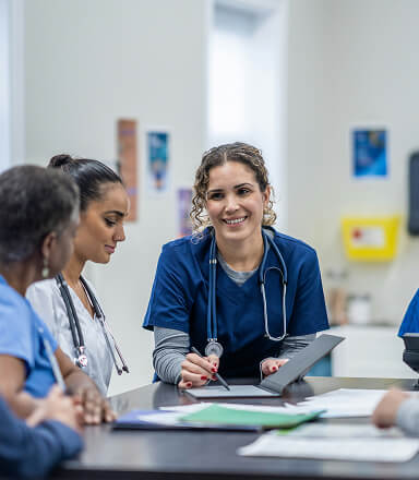 Smiling healthcare worker in navy scrubs holding a clipboard during a meeting with colleagues.