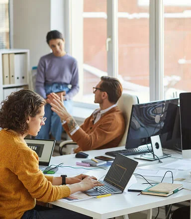 Three coworkers in an office: one typing on a laptop, two talking near a window.
