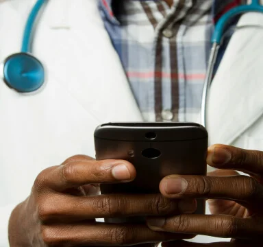 Doctor wearing a white coat and stethoscope holding and using a smartphone with both hands.