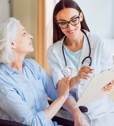 A female doctor with glasses and a stethoscope smiles while holding a clipboard and talking with an elderly female patient in a wheelchair.
