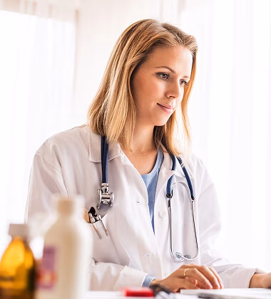 A female doctor with blonde hair wearing a white coat and stethoscope, looking down and typing on a laptop in a bright room.