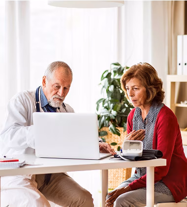 A male doctor in a white coat uses a laptop while discussing medical information with a middle-aged female patient seated across from him at a table in a well-lit room with a plant in the background.