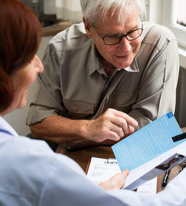 Elderly man wearing glasses reading a blue pamphlet held by a woman with reddish hair, with a document and pen on the table.