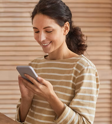 Smiling woman with dark hair tied back wearing a beige and white striped shirt, looking at and using a smartphone.