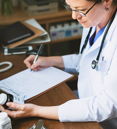 A female doctor wearing glasses and a white coat with a stethoscope around her neck writes on a clipboard using a pen while holding a prescription pill bottle in her other hand.