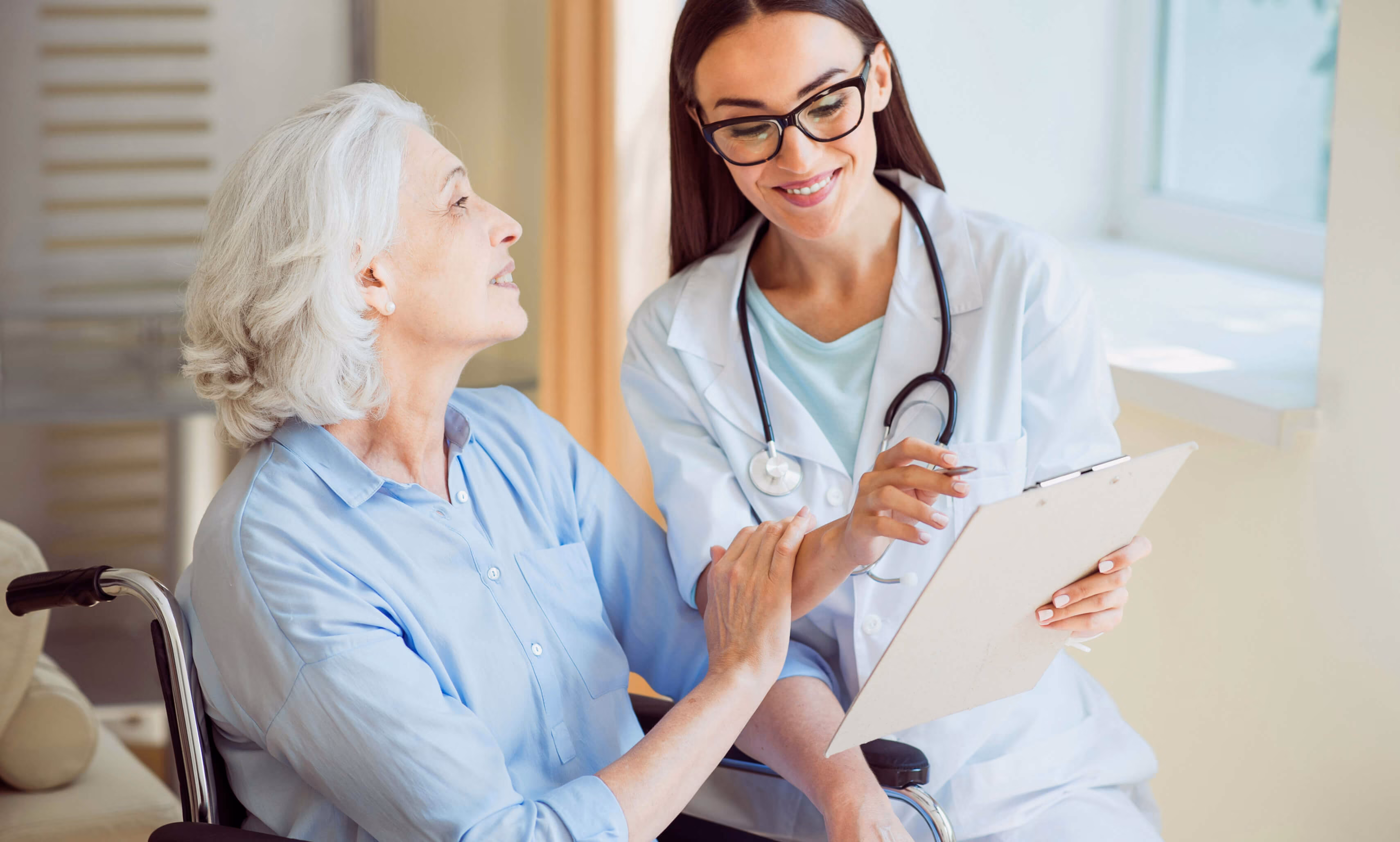 Smiling female doctor with glasses and stethoscope showing a clipboard to an elderly woman with gray hair sitting in a wheelchair.