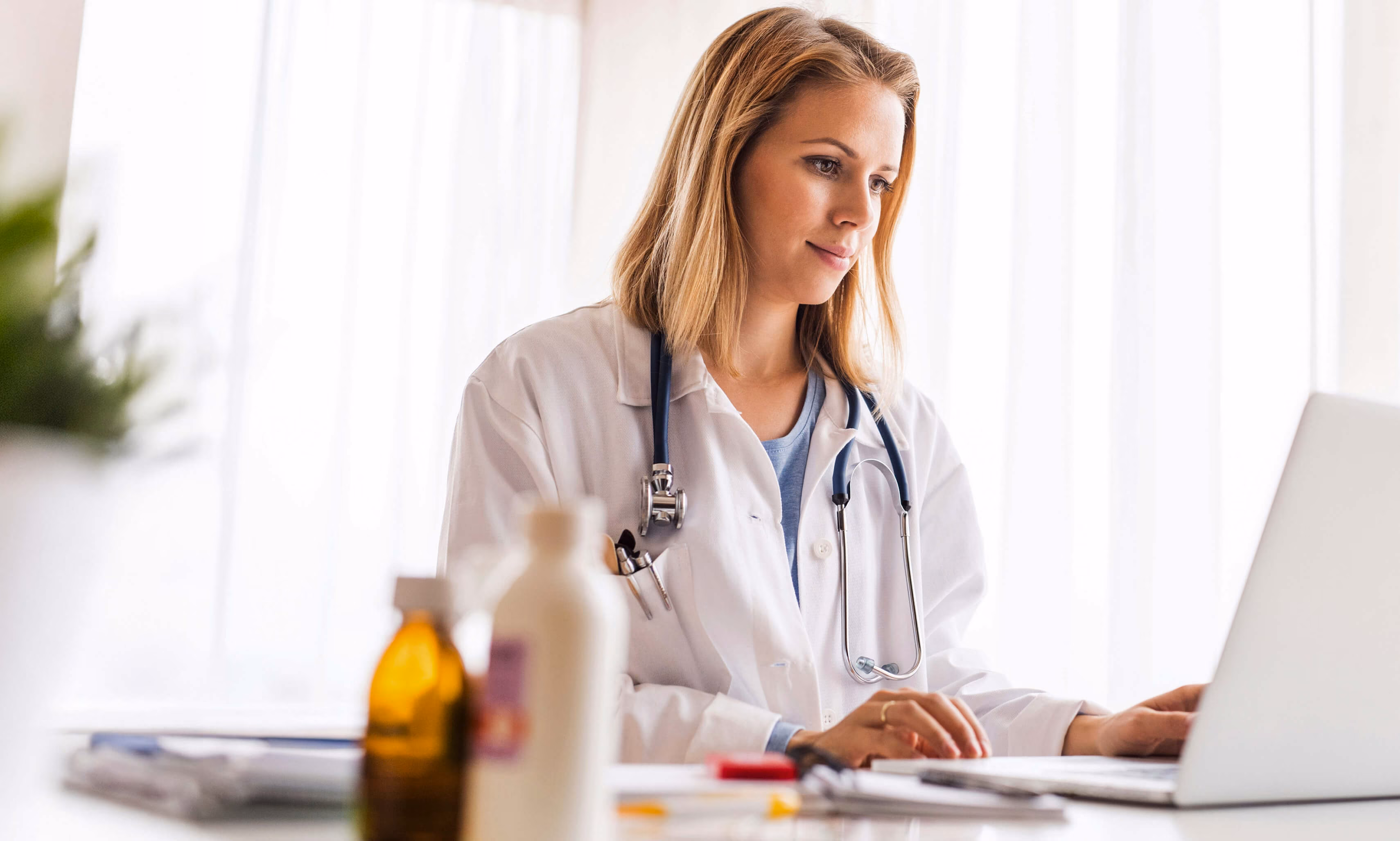 Female doctor with blonde hair wearing a white coat and stethoscope working on a laptop at a desk with medicine bottles in the foreground.