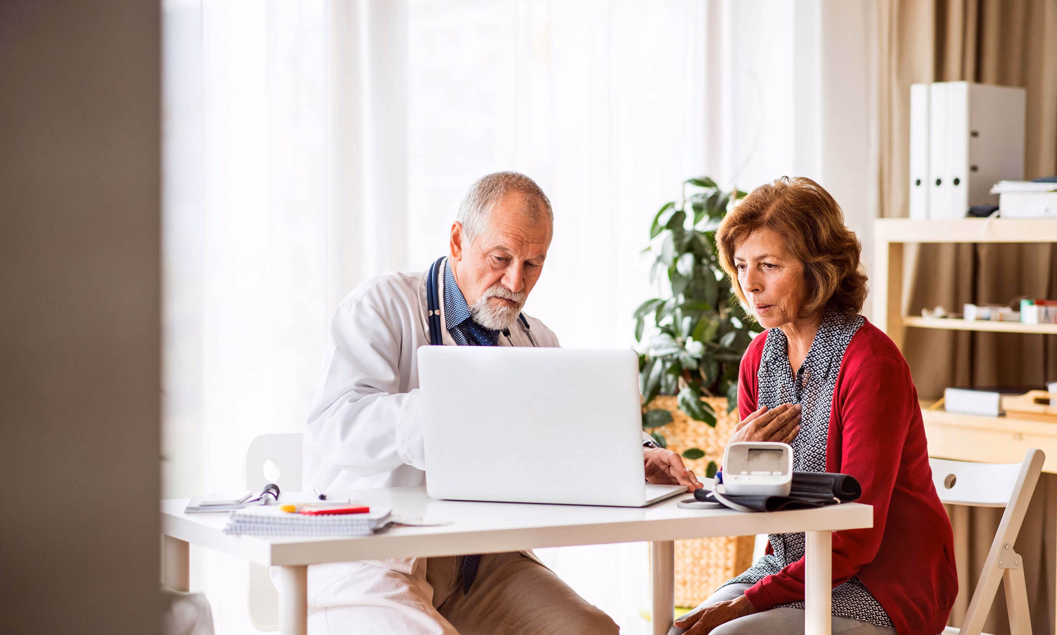 Senior doctor in a white coat consulting a middle-aged woman while sitting at a desk with a laptop and medical equipment, including a blood pressure monitor.