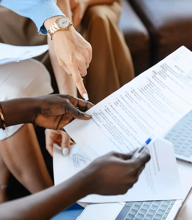 Two people reviewing and pointing at a printed document with text while holding papers over a laptop keyboard.