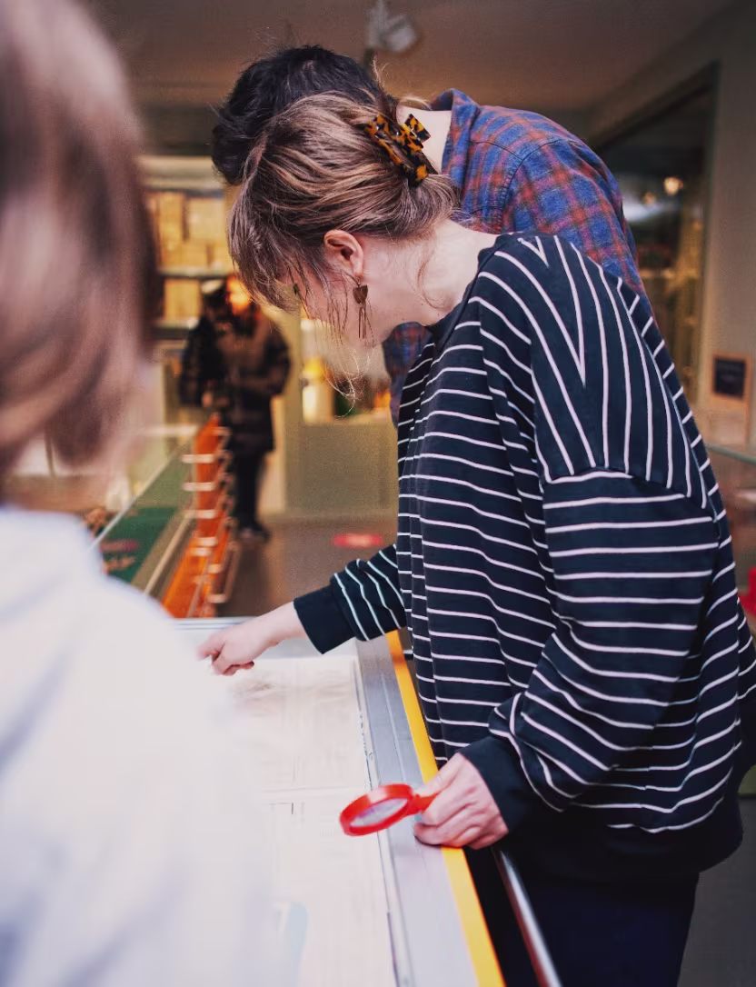Two people closely examining a display table, one holding a red magnifying glass in a public indoor setting.