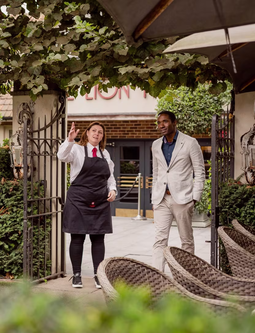Waitress in apron gestures while talking to man in suit jacket outside restaurant gate.