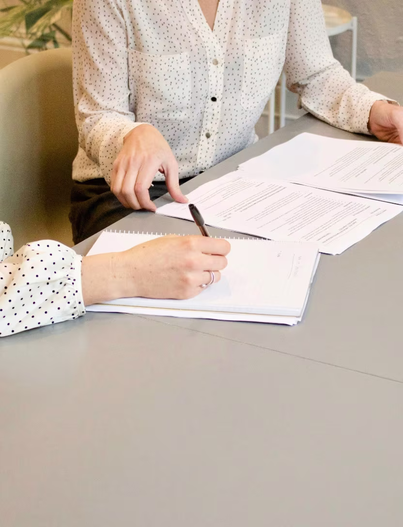 Two people at a table with one pointing at documents and the other writing on a notepad.