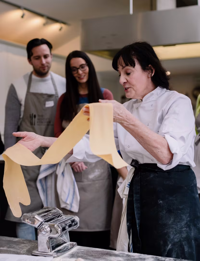 Chef demonstrating pasta dough handling to people in aprons during a cooking class.
