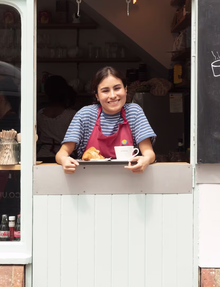 Smiling barista in apron serving croissant and coffee from a cafe window.