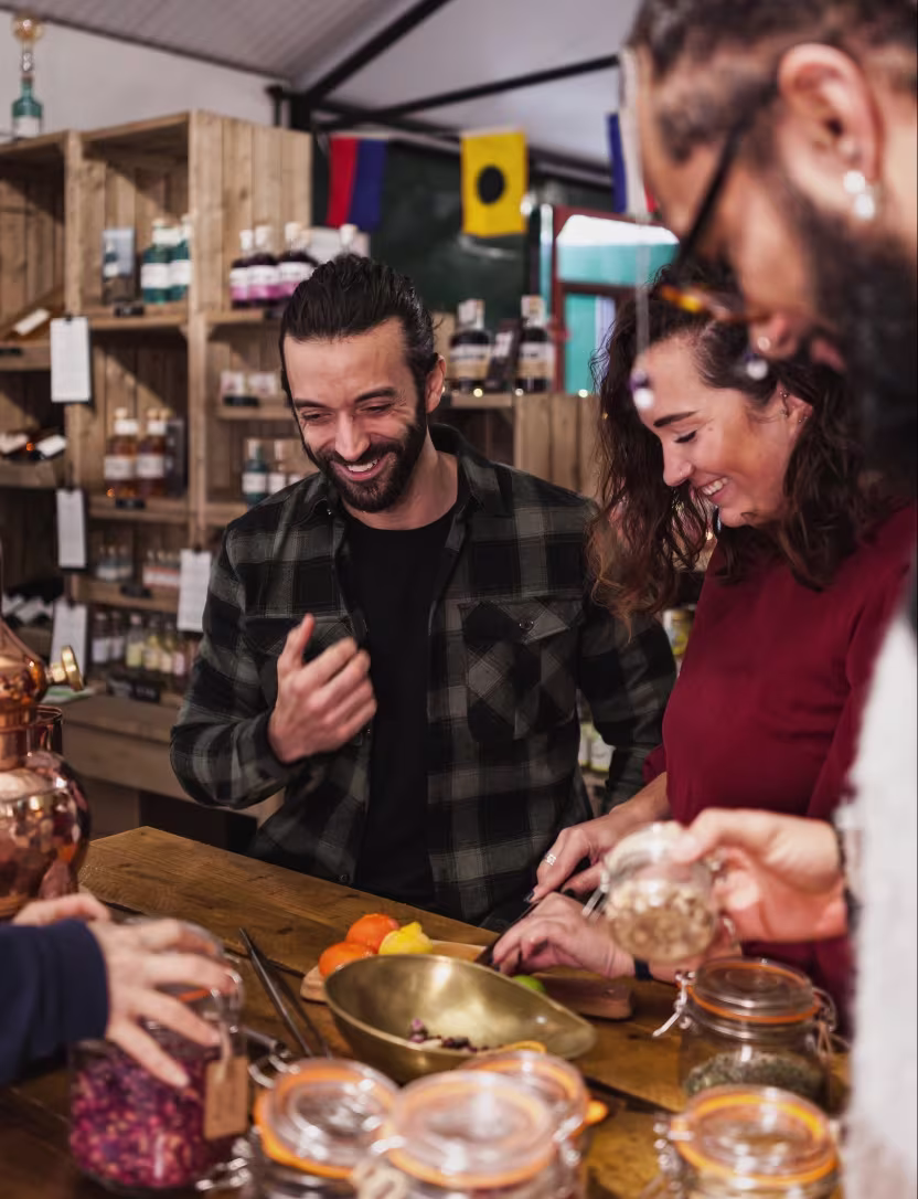 A man and woman laughing together while preparing food at a wooden table with jars and spices.