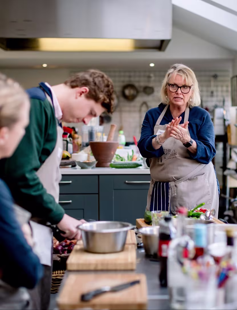 Woman in apron instructs while man prepares food in a kitchen during cooking class.