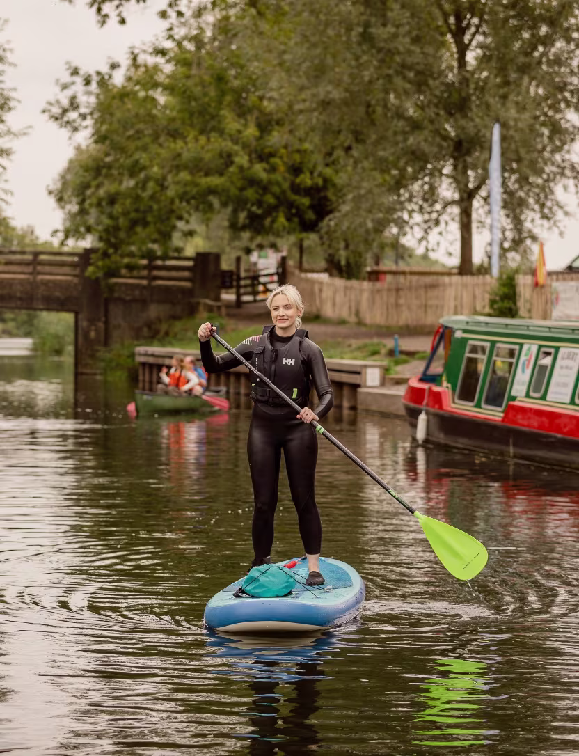 Woman in black wetsuit paddleboarding on calm canal water near a moored narrowboat and green trees.