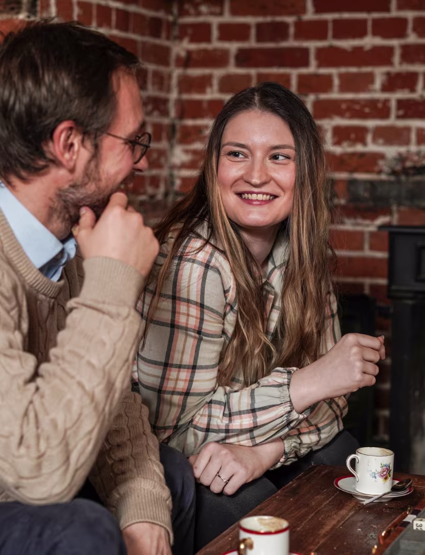 Two people sitting and smiling, enjoying coffee in a cozy setting with exposed brick walls.