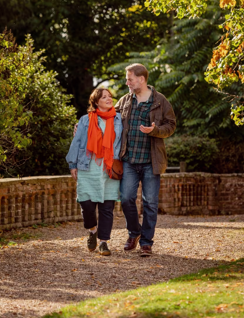 Middle-aged couple walking arm in arm on a leafy garden path under autumn sunlight.