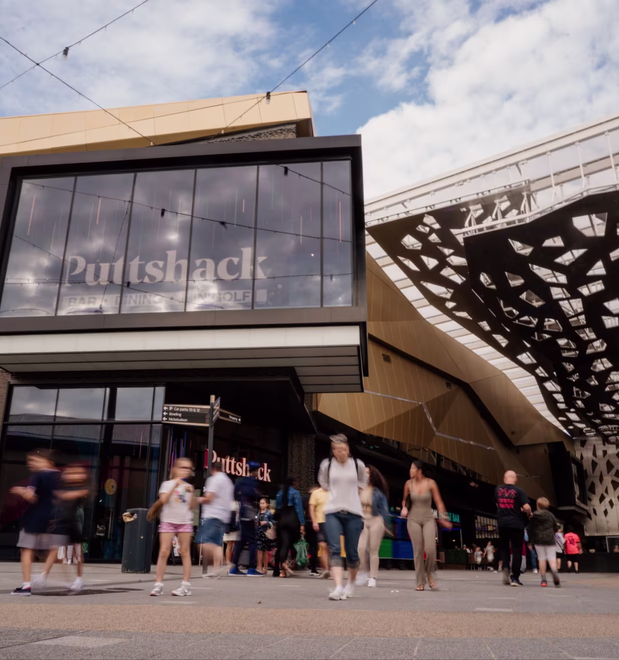 People walking past Puttshack, a modern entertainment venue with a golf bar and large glass facade.